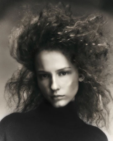 Portrait of a woman with voluminous curly hair wearing a dark turtleneck sweater against a neutral background