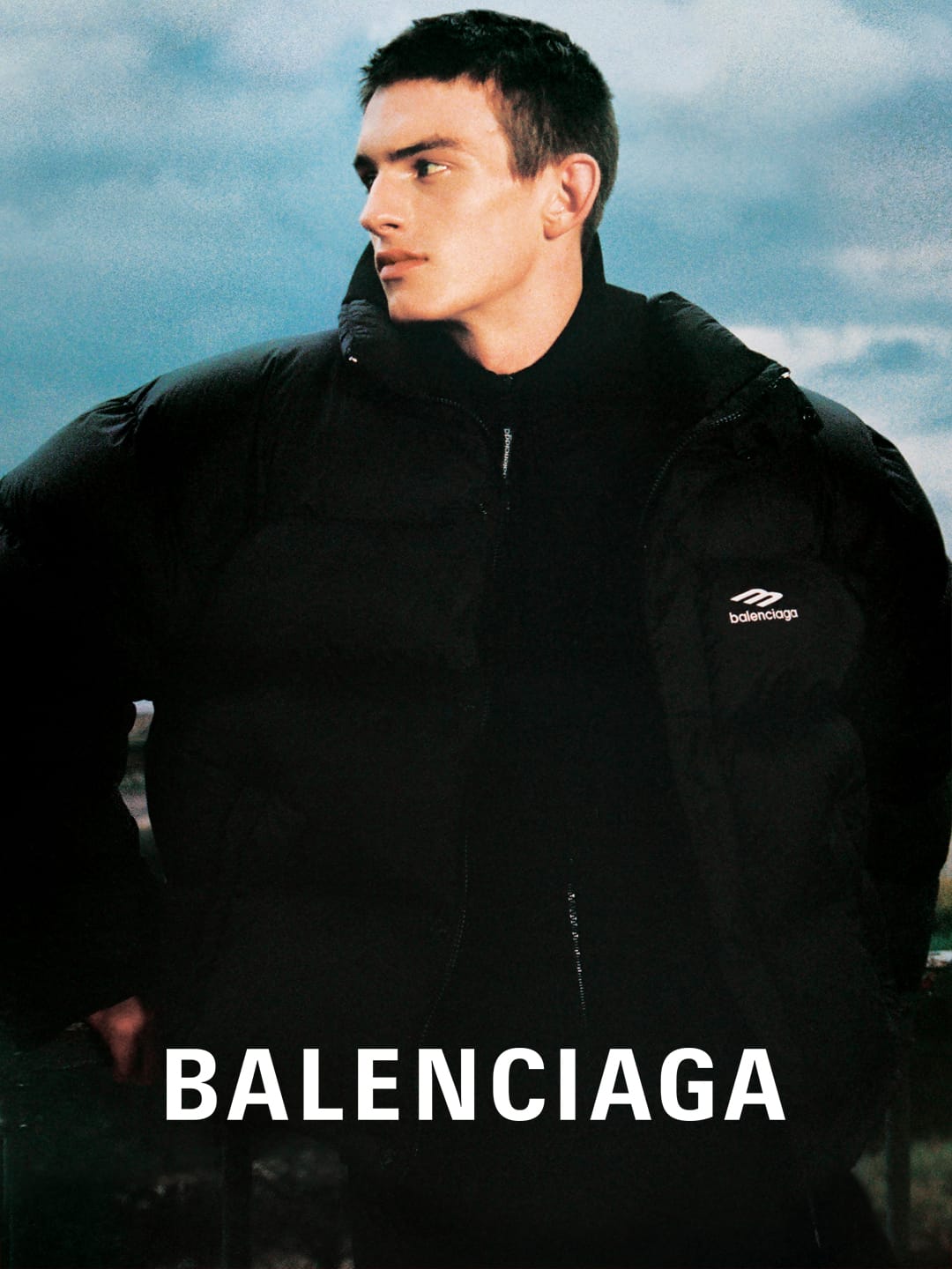 A male model poses in a black Balenciaga puffer jacket against a textured, slightly blurred background of sky and urban elements.