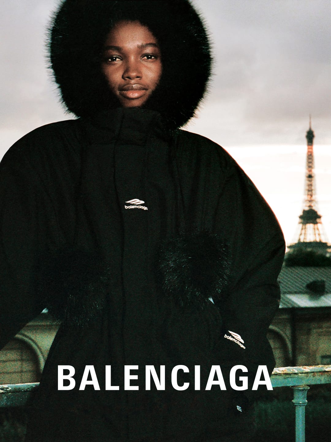 Fashion portrait of an adult Black woman in a black hooded parka with fur trim, Eiffel Tower in the background, Balenciaga branding.