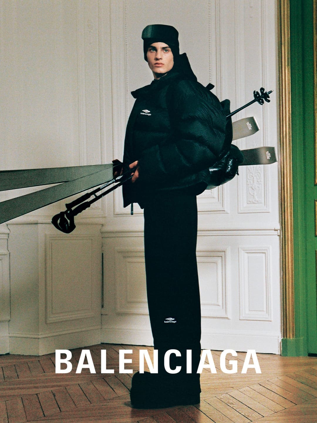Fashion shot of a male model wearing black skiwear and a hat carrying skis on his back in an ornate interior. Balenciaga logo visible.