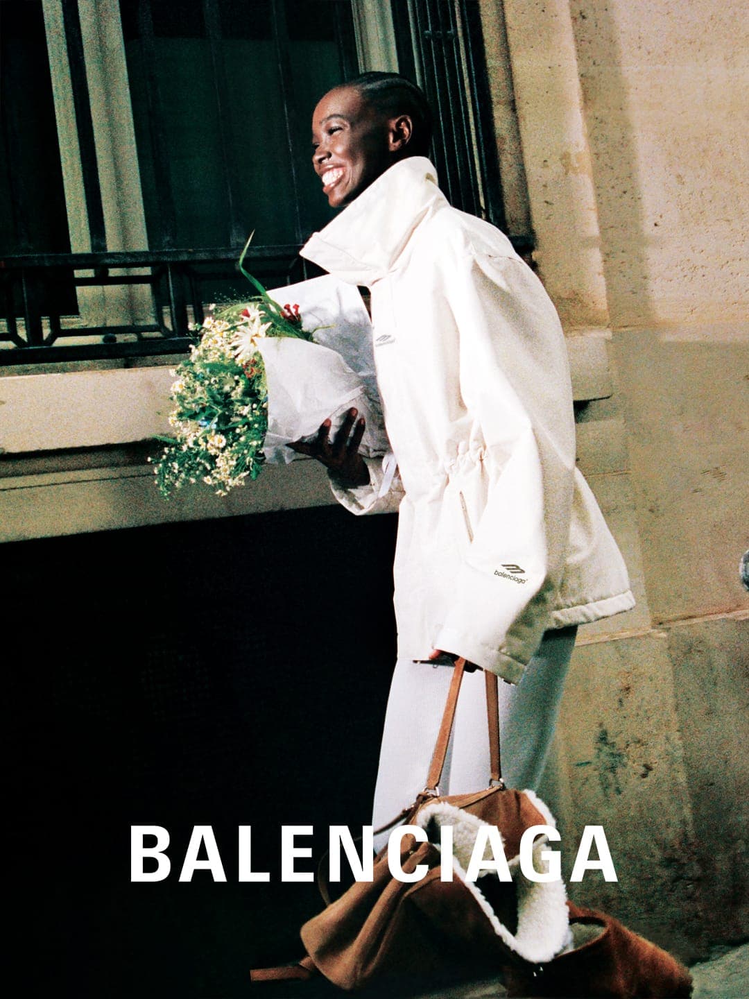 A smiling Black woman in a white jacket holds a bouquet with a Balenciaga bag, walking past a dark window in an outdoor, urban setting.