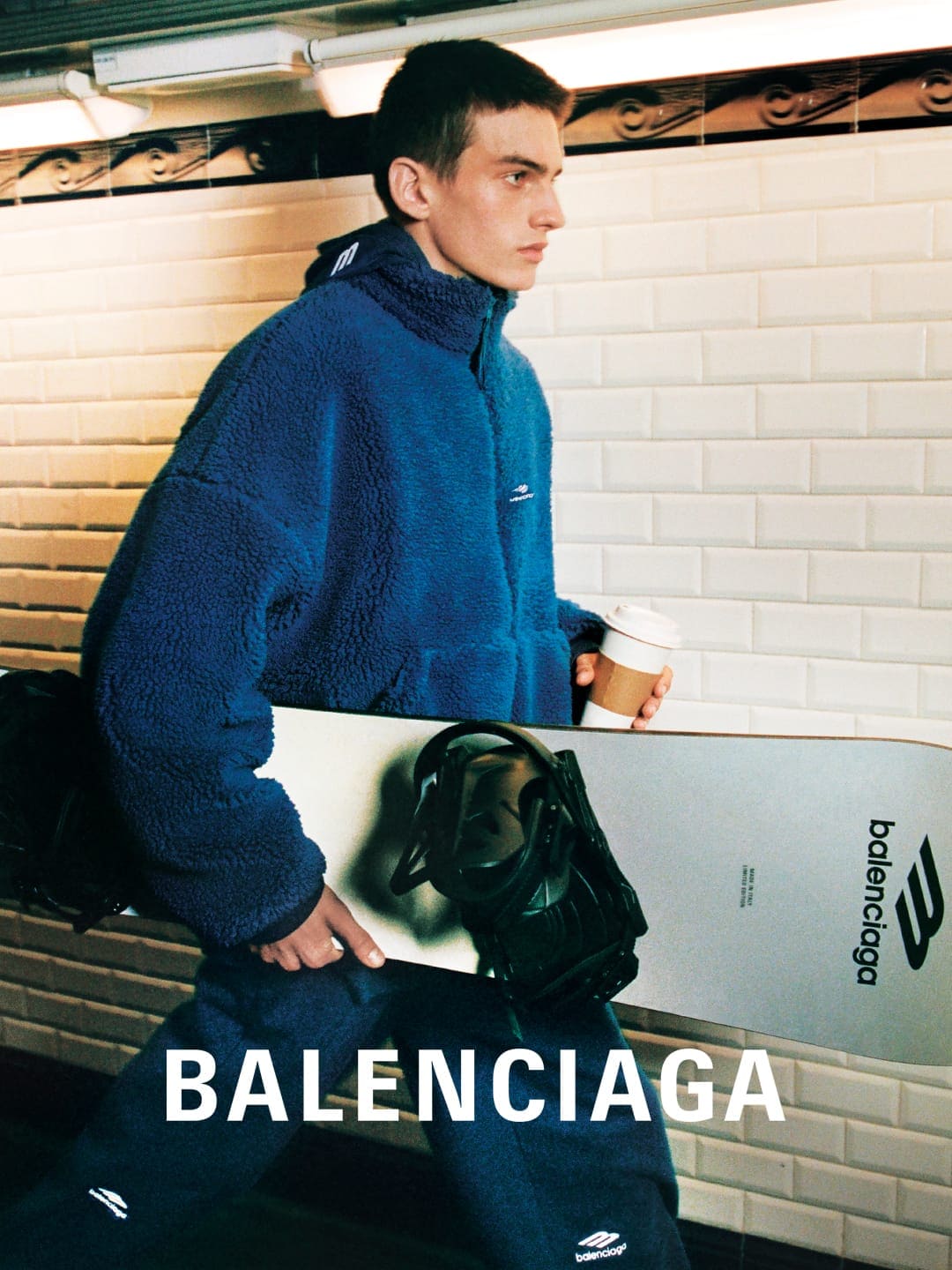 Man in blue fleece jacket holds Balenciaga snowboard indoors. Subway tiles and coffee cup add to the urban, sporty fashion scene.
