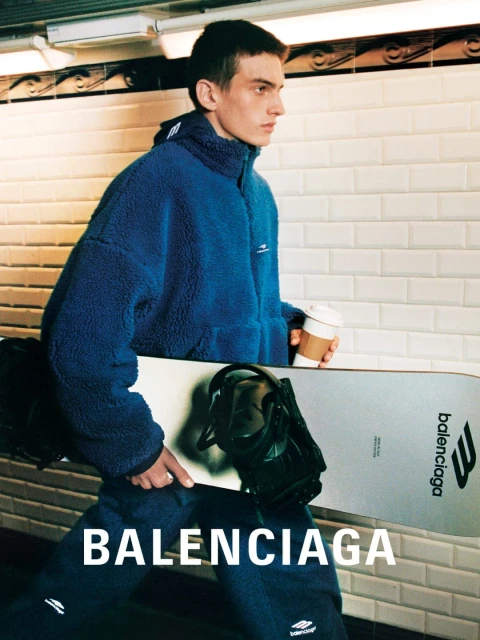 Man in blue fleece jacket holds Balenciaga snowboard indoors. Subway tiles and coffee cup add to the urban, sporty fashion scene.