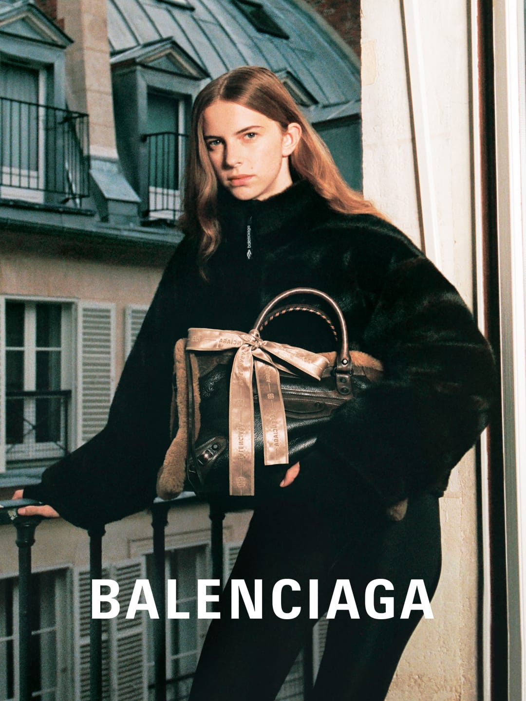 Fashion editorial shot of a young woman wearing a black jacket and holding a handbag with a building in the background.