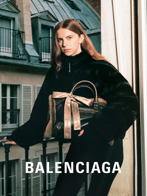 Fashion editorial shot of a young woman wearing a black jacket and holding a handbag with a building in the background.