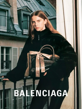 Fashion editorial shot of a young woman wearing a black jacket and holding a handbag with a building in the background.