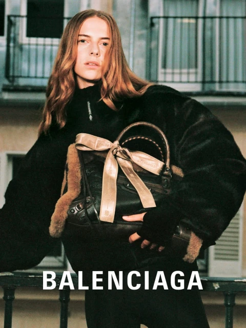 Fashion shot of a long-haired woman modeling Balenciaga with a brown bag on a balcony against a building backdrop and large brand label.