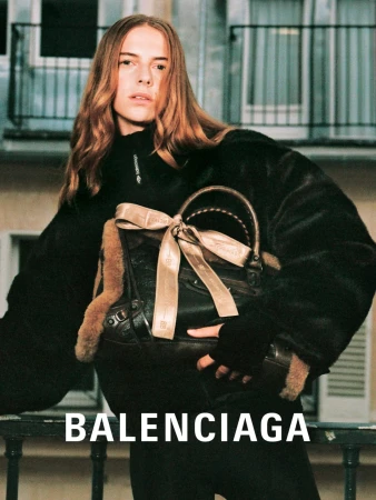 Fashion shot of a long-haired woman modeling Balenciaga with a brown bag on a balcony against a building backdrop and large brand label.