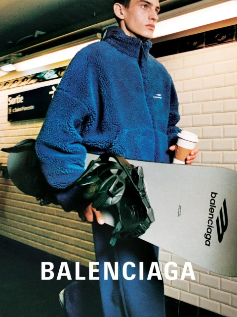 A fair-skinned man with dark hair holds a snowboard in an underground metro station, wearing a blue sherpa jacket and matching pants.