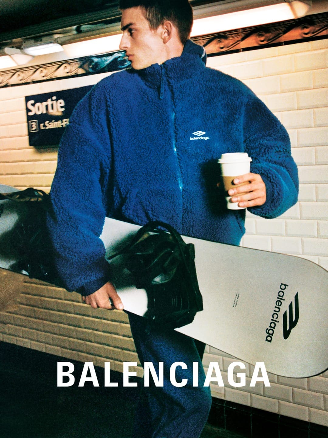 A man in a blue fleece jacket holds a Balenciaga snowboard and a coffee cup in a subway station, lit with natural light.