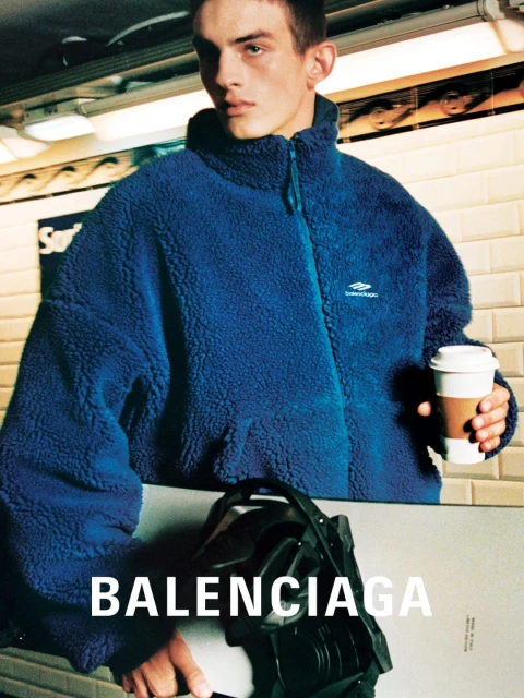 A fair-skinned man in a blue fleece jacket holds a snowboard and a coffee cup in a subway station with tan tile walls.