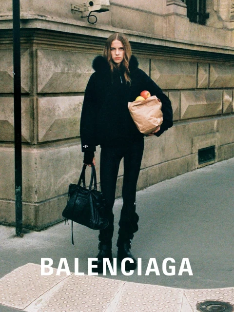 Fashion shot of woman in black outfit carrying bag and groceries walking on city street next to stone building with Balenciaga logo.