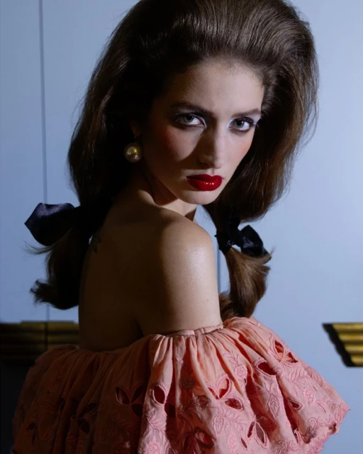 Portrait of a woman with high volume hair, wearing a peach off-the-shoulder dress with cutouts, red lipstick, and pearl earrings against a blue backdrop.