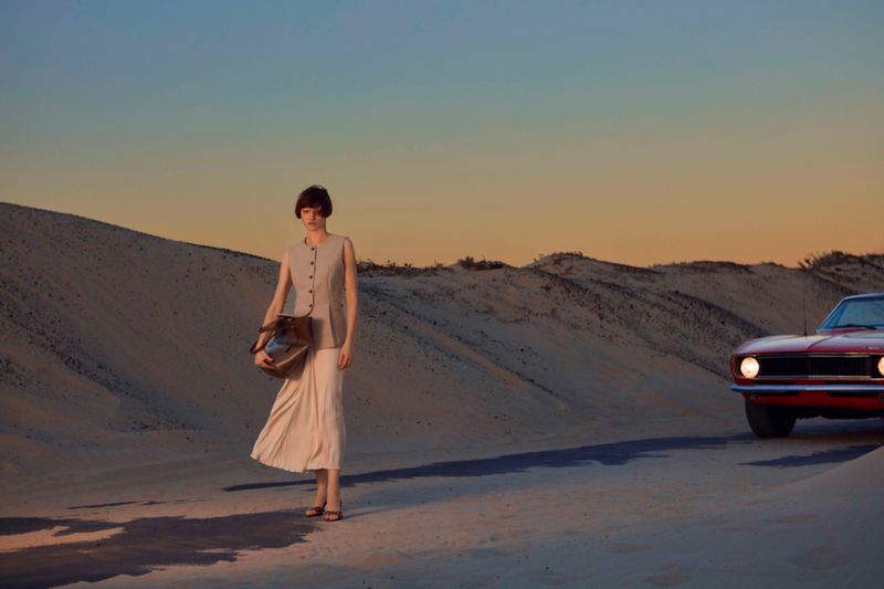 A woman poses with a handbag on a sandy landscape near a vintage red car at sunset, creating a blend of fashion and automotive.
