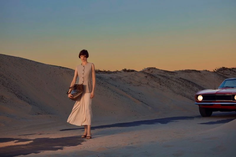 A woman poses with a handbag on a sandy landscape near a vintage red car at sunset, creating a blend of fashion and automotive.