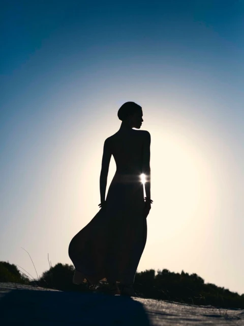 Silhouette of an adult female in a flowing dress standing on a sandy surface against a bright sky, evoking nature and fashion.