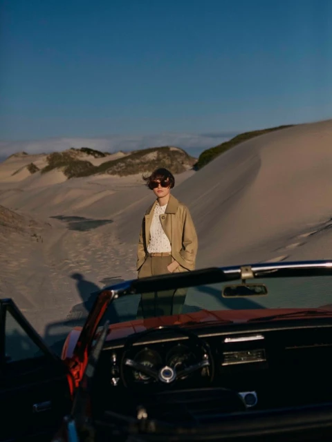 A woman in a tan coat and sunglasses stands near a classic convertible car on a sandy beach with dunes under a clear blue sky.