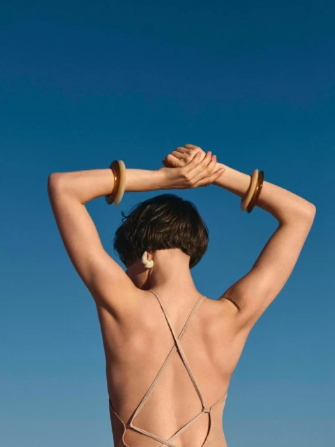 Back view of a woman in a bathing suit with bracelets on her raised arms against a clear blue sky, bathed in bright sunlight.