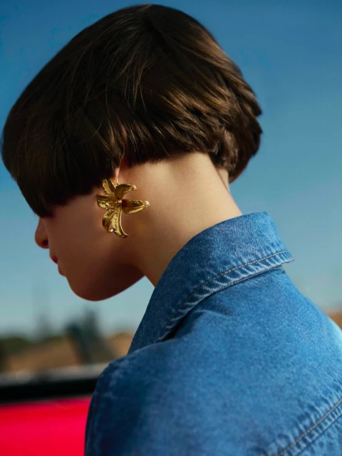 Woman in a denim jacket wearing a large, gold flower-shaped earring against a gradient blue and brown background, close-up shot.