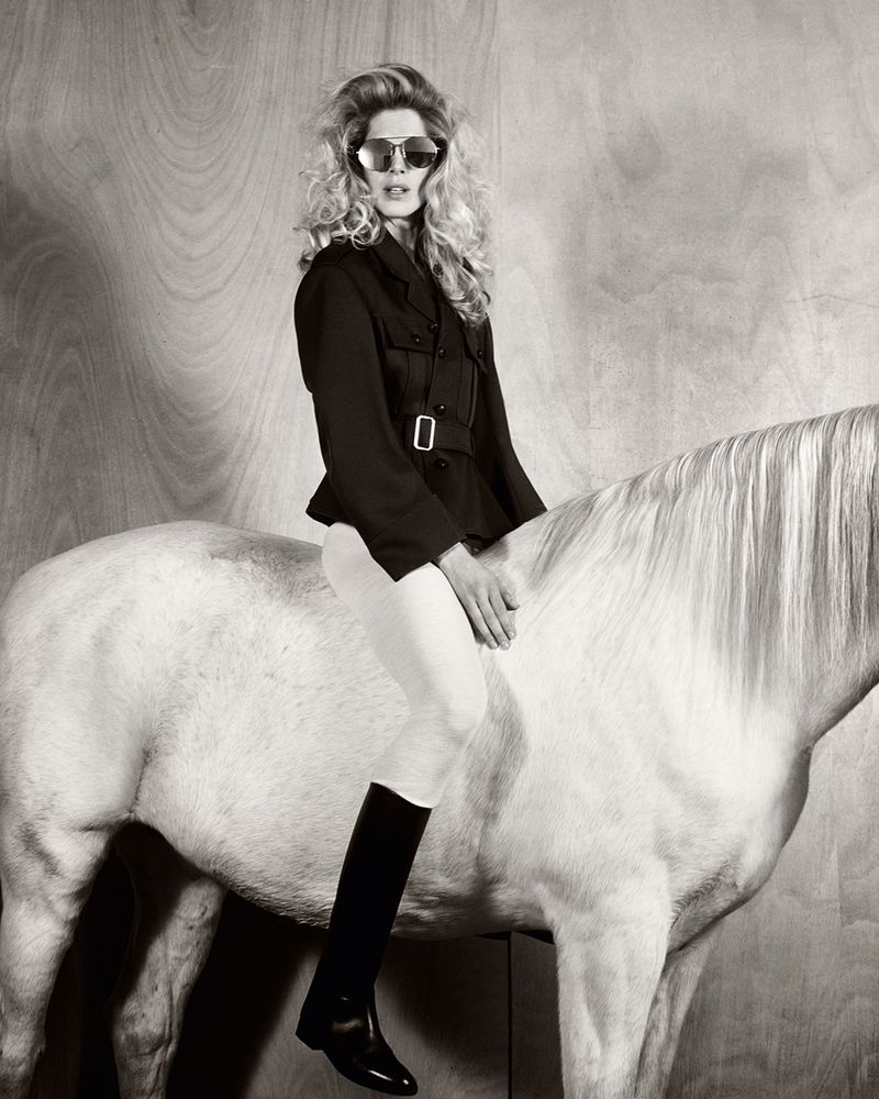 Monochrome fashion shot of a woman in sunglasses, a black jacket, and riding boots, sitting atop a white horse against a wood panel.