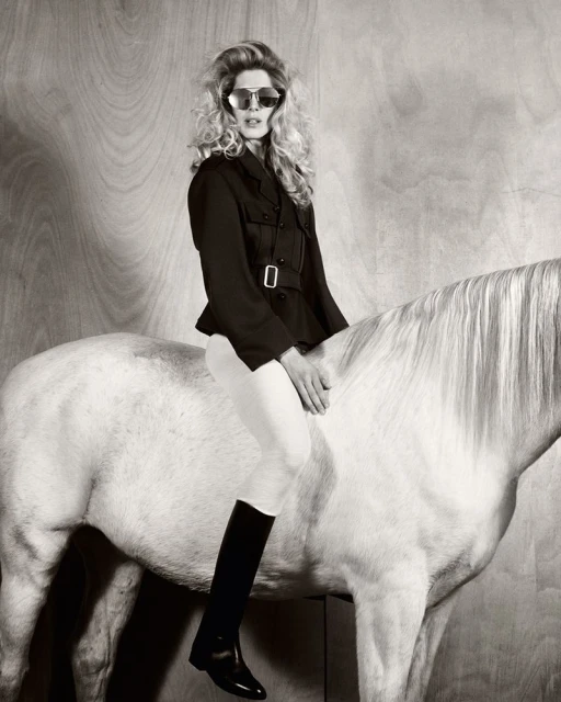 Monochrome fashion shot of a woman in sunglasses, a black jacket, and riding boots, sitting atop a white horse against a wood panel.