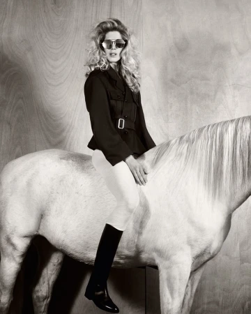 Monochrome fashion shot of a woman in sunglasses, a black jacket, and riding boots, sitting atop a white horse against a wood panel.