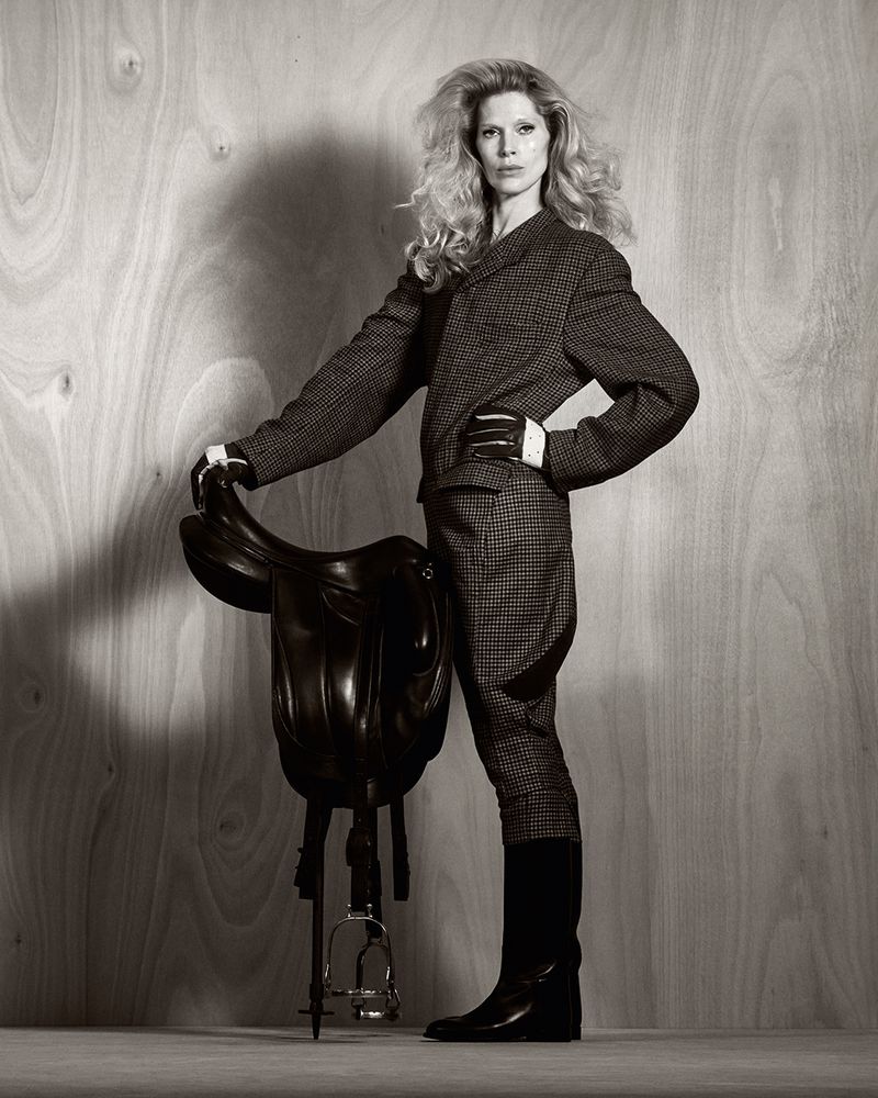 Monochrome fashion shot of an adult woman in a blazer and jodhpurs, posing with a black horse saddle against a wood-panel backdrop.