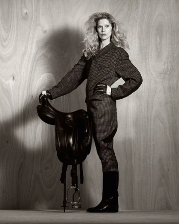 Monochrome fashion shot of an adult woman in a blazer and jodhpurs, posing with a black horse saddle against a wood-panel backdrop.