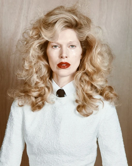 Close-up portrait of a blonde woman with curly hair, wearing a white textured top and a dark tie against a neutral backdrop.