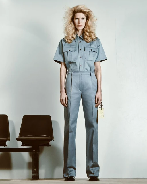 A female fashion model stands in a matching light blue shirt and pants in a studio, with black plastic chairs in the foreground.