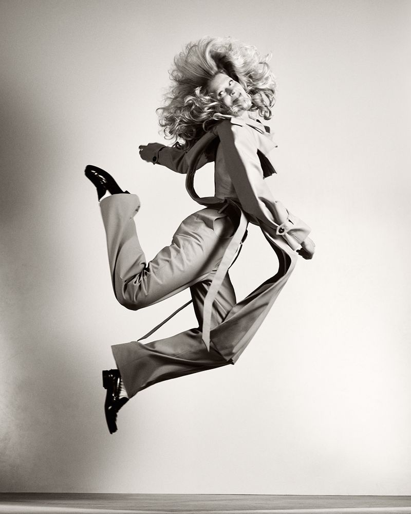 Black and white studio shot of a blonde adult woman in a coat and pants jumping in the air with one leg up and her arm outstretched.