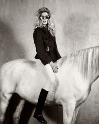 Iselin Steiro sitting on a white horse wearing a dark jacket, light trousers, tall boots and sunglasses against a wood panel wall