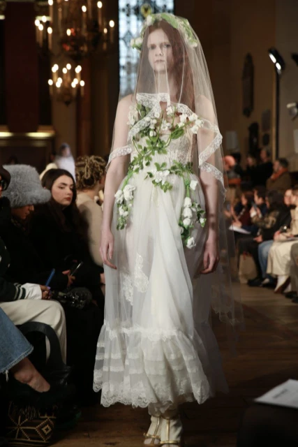 Runway model in a floral white dress and veil walks the runway during a fashion show, captured in a medium shot.