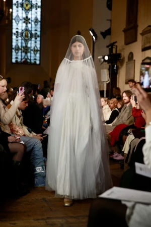 Runway fashion show featuring a model in a white dress and veil walking past a seated audience during an indoor event.