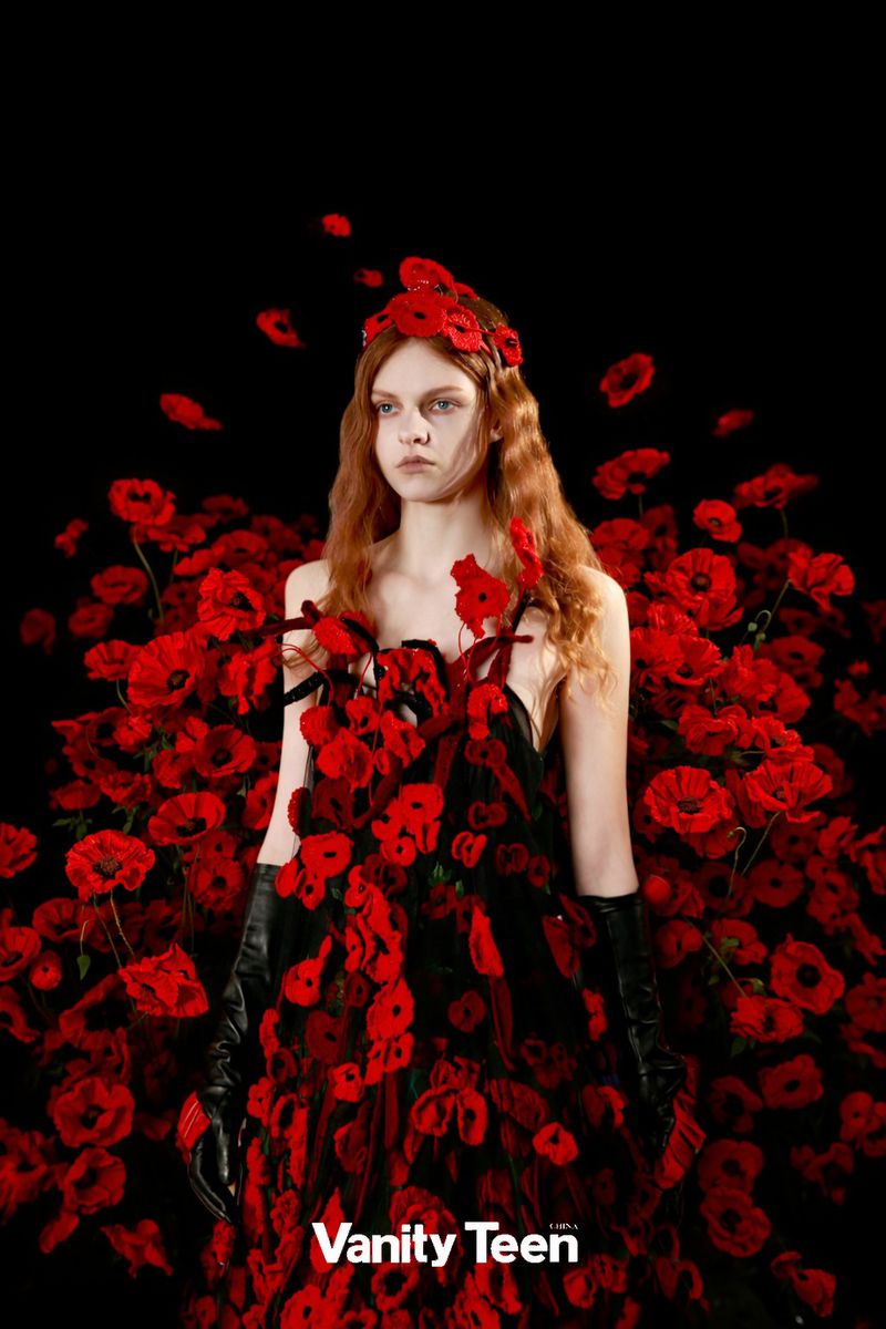 Portrait of a young woman in a floral dress and headpiece posing in front of a black background with bright red poppy flowers.