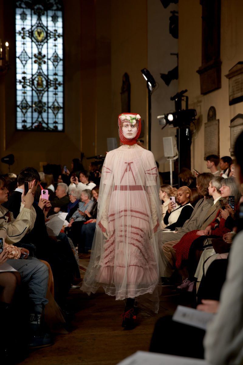 Fashion runway show featuring a model in a pink dress with floral headpiece, surrounded by a seated audience in a hall with large windows.