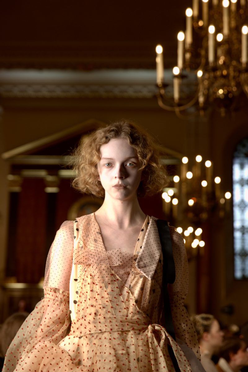Young woman in a peach-colored polka dot dress stands in an ornate interior with chandeliers, showcasing a fashion or beauty aesthetic.
