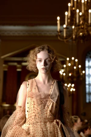 Young woman in a peach-colored polka dot dress stands in an ornate interior with chandeliers, showcasing a fashion or beauty aesthetic.