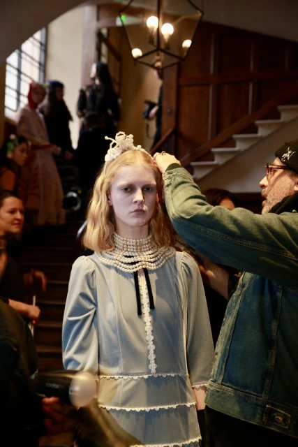 Model getting hair adjusted backstage at fashion show. Young woman in dress with pearl collar, ornate headpiece, stairs in background.