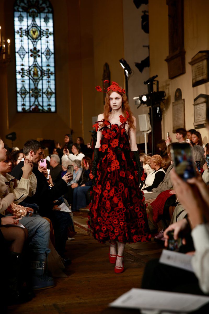 Runway show features a model in a striking red floral gown, with an audience watching in a church-like setting with stained glass windows.