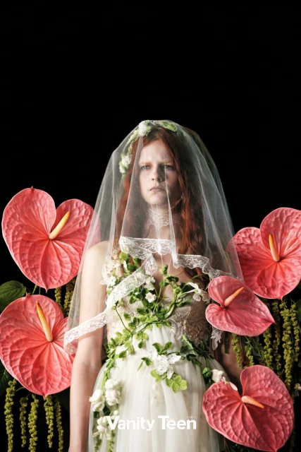 Portrait of a red-haired woman wearing a floral-covered dress and a veil, framed by heart-shaped flowers against a black background.