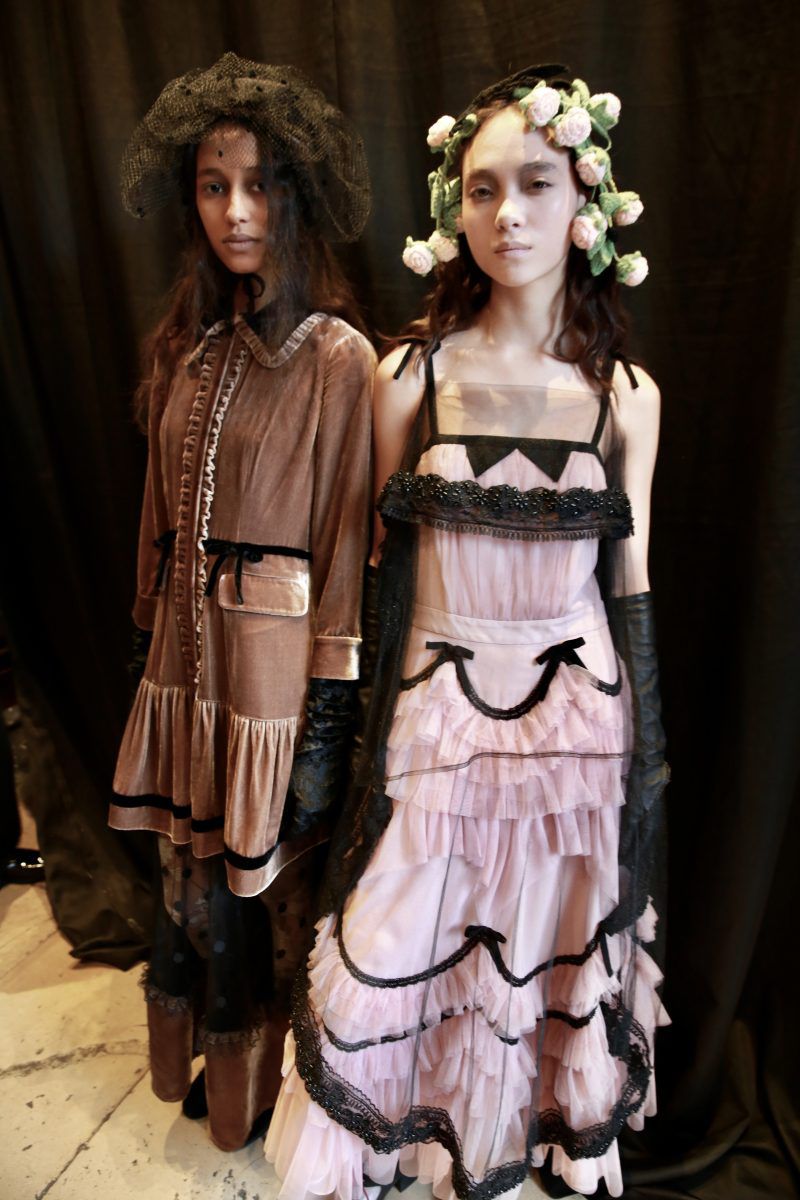 Two female fashion models pose in elaborate vintage-style dresses and floral crowns against a dark backdrop in a studio setting.
