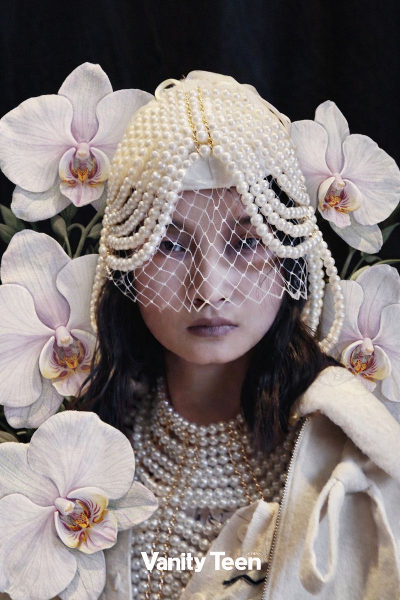Young woman with dark hair wears a pearl headpiece with netting and a pearl necklace, surrounded by white orchids against a black background.