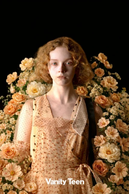 Portrait of a young woman with curly red hair in a floral dress surrounded by peach and white roses against a dark background.