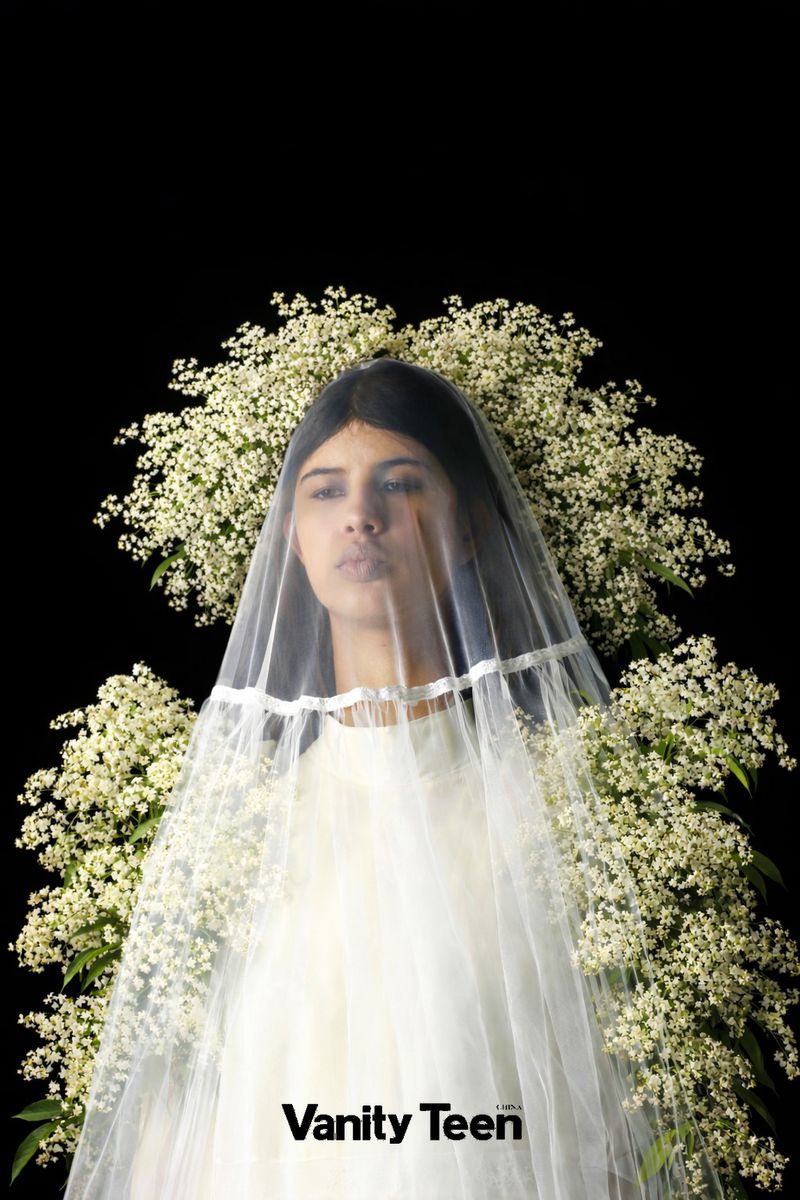 Woman in sheer veil surrounded by white flowers against black backdrop for Vanity Teen editorial, soft lighting, beauty-focused, portrait orientation.