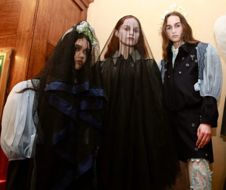 Three models pose indoors wearing black dresses with veils. One model wears a black jacket with light blue sleeves and floral tights.