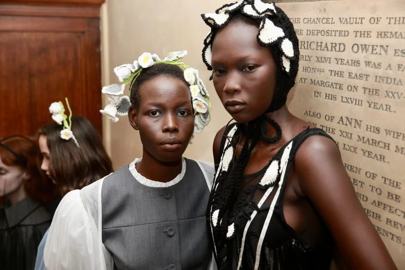 Two African American fashion models in headwear and elegant clothing strike poses against a textured wall inside, showcasing high fashion.
