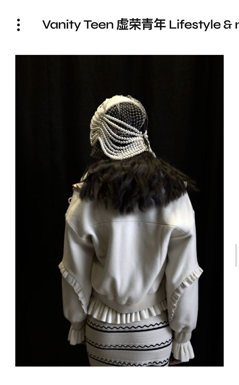 Back view of a fashion model in an ornate pearl headpiece and ruffled jacket and skirt, posing against a dark backdrop.