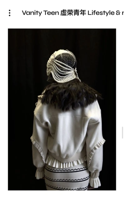 Back view of a fashion model in an ornate pearl headpiece and ruffled jacket and skirt, posing against a dark backdrop.