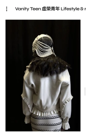 Back view of a fashion model in an ornate pearl headpiece and ruffled jacket and skirt, posing against a dark backdrop.